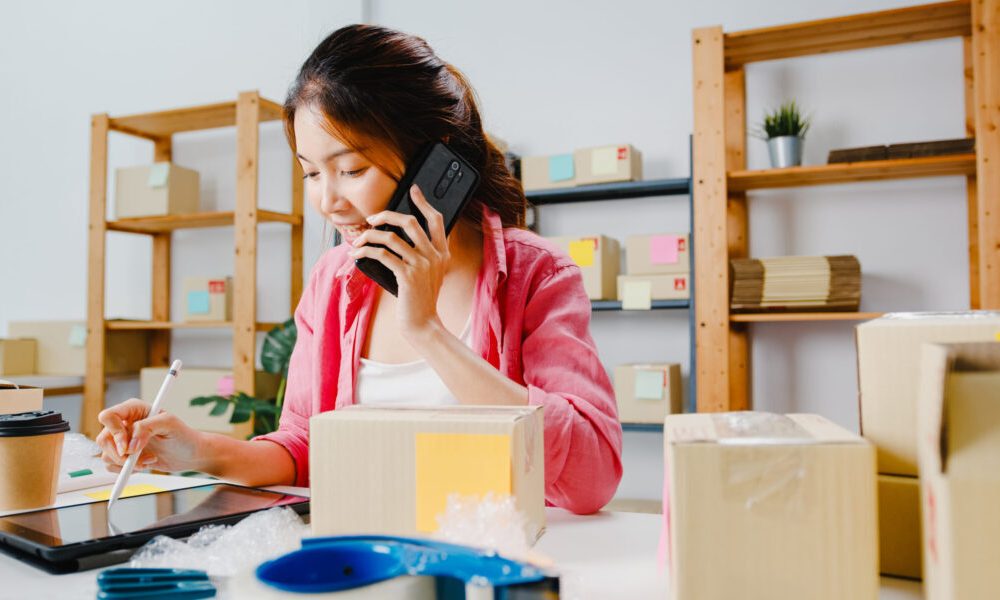 Young Asia businesswoman using mobile phone call receiving purchase order and check product on stock, work at home office. Small business owner, online market delivery, lifestyle freelance concept.